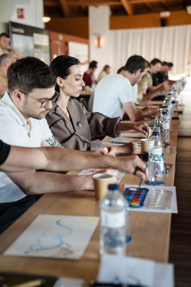 Eine lange Tafel, an der Teilnehmende konzentriert mit Wasserfarben malen